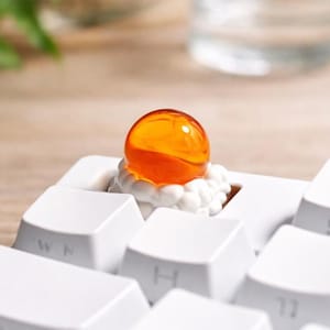 May include: A custom keycap featuring an orange, spherical design atop a white cloud-like base. The keycap is set on a white keyboard, with other keys visible in the foreground. The keycap is a unique accessory for mechanical keyboards.