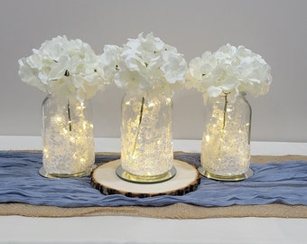May include: Three clear glass jars with white hydrangea flowers and white fairy lights inside. The jars are sitting on a wooden slice and a blue and brown runner.