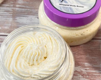 Pode incluir: Dois frascos transparentes de manteiga corporal batida. Um frasco está aberto, revelando um redemoinho de produto cremoso esbranquiçado. O outro frasco tem uma tampa roxa com o texto "Aroma Nova Melis Frost Whipped Body Butter". Os frascos estão sobre uma superfície de madeira marrom claro.