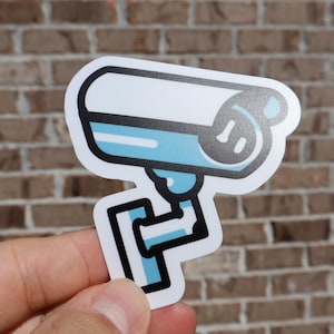 May include: A white and blue sticker of a cartoon security camera with a black outline.