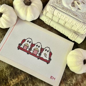 May include: A white needlepoint canvas featuring a cross-stitch design of three white ghosts sitting in a row on a red theater seat, each holding a striped popcorn bucket. The initials EM are stitched in red in the bottom right corner of the canvas.