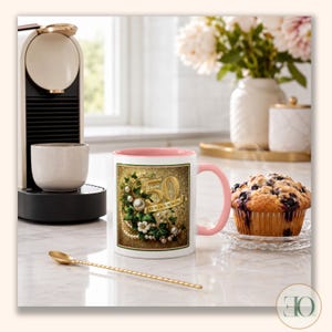 May include: A white ceramic mug with a pink interior and handle, featuring a graphic of the number 50, the text GOLDEN SOROR, green ivy leaves, and white pearls on a gold-toned background, positioned next to a blueberry muffin and a gold spoon.