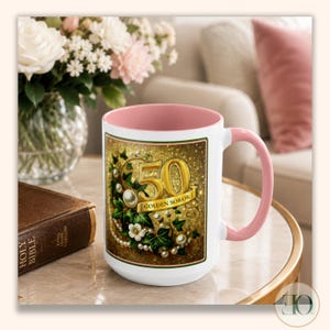 May include: A white ceramic mug with a soft pink interior and handle. The exterior features a rectangular graphic with a gold number 50, the text GOLDEN SOROR, green ivy leaves, white flowers, and strings of white pearls on a gold glitter background.