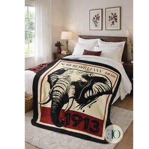 May include: A black, red, and cream-colored blanket featuring a large elephant graphic. The blanket has the text "SOROR BRITTANY Fall 2025" and "1913". The blanket is draped over a bed with white bedding.