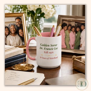 May include: A white ceramic mug with a pink handle and interior, featuring the text Golden Soror Dr. Francie Lee, Fall 1976, and 50 Years of Sisterhood in green and red lettering. The mug contains three pens and sits on a desk with office supplies.