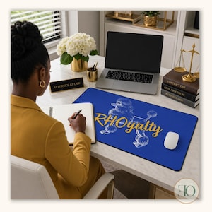 May include: A rectangular blue desk mat featuring a white line-art illustration of a poodle and the gold-colored text RHOyalty printed across the center. The mat is positioned on a white desk surface next to a laptop, a computer mouse, and books.