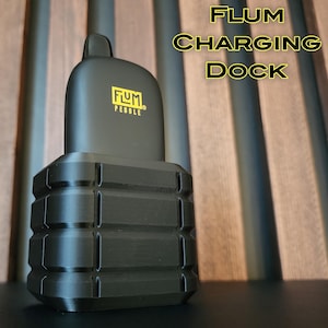 May include: A black charging dock for a FLUM Pebble vape device. The dock has a textured, ridged design and the vape device is sitting in the dock.