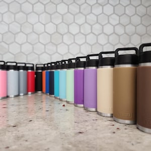 May include: A collection of insulated water bottles in various colors, including pink, gray, red, blue, and brown. Each bottle has a black handle and lid, with a silver base. The bottles are arranged in a row on a light-colored surface.