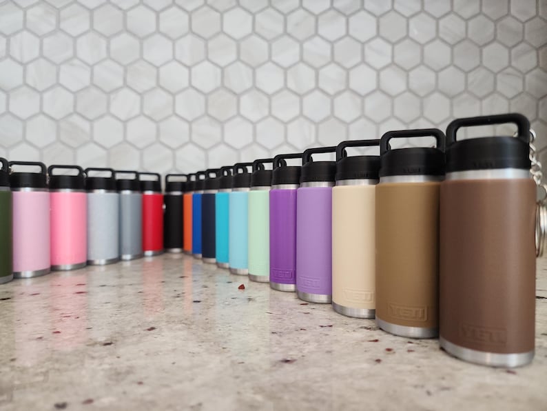 May include: A collection of insulated water bottles in various colors, including pink, gray, red, blue, and brown. Each bottle has a black handle and lid, with a silver base. The bottles are arranged in a row on a light-colored surface.