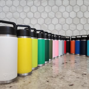 May include: A collection of insulated water bottles in various colors, including white, yellow, green, pink, and blue. Each bottle has a black handle and lid, with a silver base. The bottles are arranged in a row on a countertop.