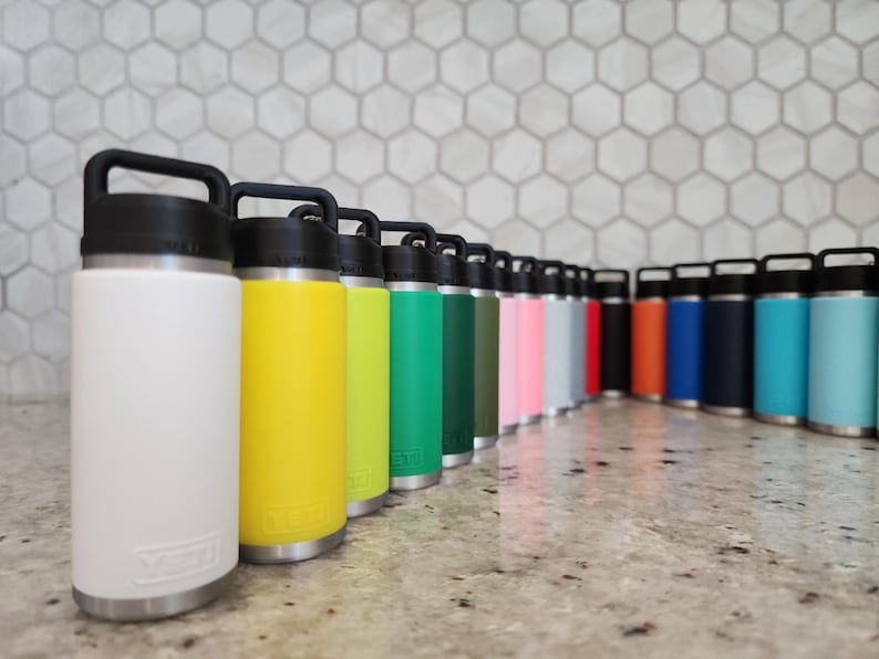 May include: A collection of insulated water bottles in various colors, including white, yellow, green, pink, and blue. Each bottle has a black handle and lid, with a silver base. The bottles are arranged in a row on a countertop.