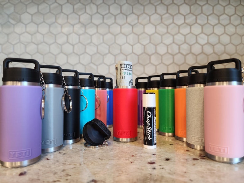 May include: A collection of insulated water bottles in various colors, including purple, gray, black, teal, pink, red, yellow, green, and orange. Each bottle has a black handle and lid. A ChapStick and a rolled-up $100 bill are also visible.