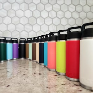 May include: A collection of insulated water bottles in various colors, including purple, pink, green, blue, black, and white. Each bottle has a black handle and lid, with a silver base. The bottles are arranged in a row on a reflective surface.