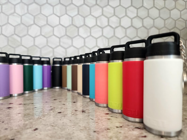 May include: A collection of insulated water bottles in various colors, including purple, pink, green, blue, black, and white. Each bottle has a black handle and lid, with a silver base. The bottles are arranged in a row on a reflective surface.