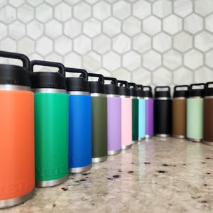 May include: A collection of insulated water bottles in various colors, including orange, green, blue, and purple. Each bottle has a black handle and lid, with a silver base. The bottles are arranged in a row on a countertop.