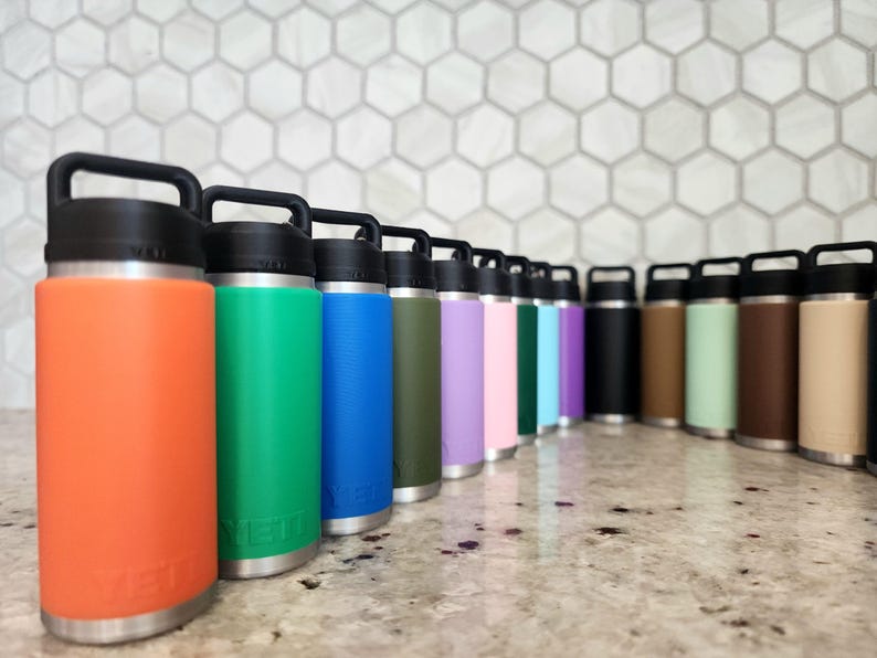 May include: A collection of insulated water bottles in various colors, including orange, green, blue, and purple. Each bottle has a black handle and lid, with a silver base. The bottles are arranged in a row on a countertop.