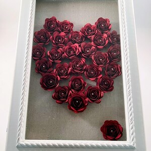 May include: A white frame with a rope design border encloses a heart shape made of burgundy paper roses. The roses are arranged in a heart shape on a gray fabric background.
