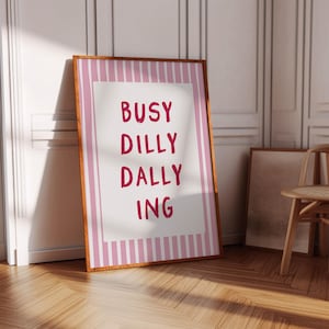 以下が含まれることがあります： 白い中央パネルに濃いピンク色の文字で「BUSY DILLY DALLY ING」と書かれた長方形の額入りウォールアート。縁取りは淡いピンクと白の縦縞模様で、薄い木製のフレームに収められています。