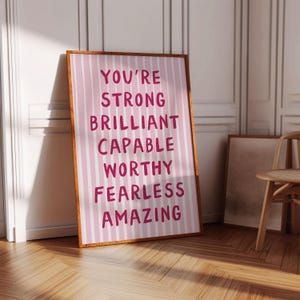 以下が含まれることがあります： ピンクと白のストライプの背景に、「You're Strong Brilliant Capable Worthy Fearless Amazing」という言葉が太字の濃いピンク色のフォントで書かれた額入りのプリント。 フレームは温かみのある茶色です。