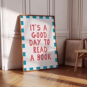Könnte beinhalten: Ein gerahmtes Poster mit dem Text "IT'S A GOOD DAY TO READ A BOOK" in roten Buchstaben. Das Poster hat einen weißen Hintergrund mit einem blau-weißen Rand und einem braunen Holzrahmen. Das Poster lehnt an einer weißen Wand.
