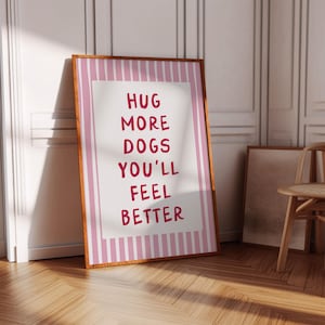 Op de afbeelding: Een ingelijste print met de tekst "HUG MORE DOGS YOU'LL FEEL BETTER" in rode letters. De print heeft een witte achtergrond met roze verticale strepen en een bruine houten lijst. De print leunt tegen een witte deur.