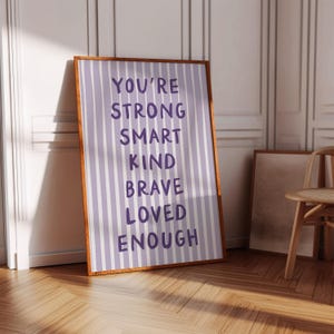 May include: A framed print with a light purple and white striped background. The print features the words "YOU'RE STRONG SMART KIND BRAVE LOVED ENOUGH" in dark purple lettering. The frame is a warm brown colour.