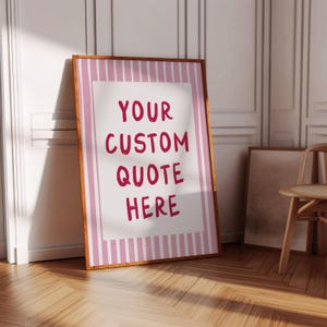 Peut inclure: Une affiche encadrée avec le texte "YOUR CUSTOM QUOTE HERE" en rouge. L'affiche a un fond blanc avec une bordure à rayures roses et un cadre en bois. L'affiche est appuyée contre un mur blanc sur un sol en bois.