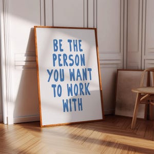 Può includere: Una stampa incorniciata con la scritta "BE THE PERSON YOU WANT TO WORK WITH" in lettere blu. La cornice è di un caldo colore marrone e la stampa è appoggiata a un muro. Una sedia di legno e un'altra stampa incorniciata sono sullo sfondo.