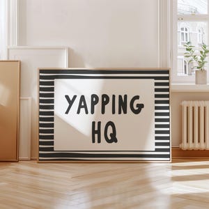 Könnte beinhalten: Ein gerahmter Kunstdruck mit dem Text "YAPPING HQ" in schwarzen, fetten Buchstaben auf weißem Hintergrund. Der Rahmen hat einen schwarz-weiß gestreiften Rand. Das Kunstwerk wird auf einem Holzboden präsentiert.