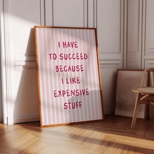 May include: A framed poster with a pink and white striped background and the text "I HAVE TO SUCCEED BECAUSE I LIKE EXPENSIVE STUFF" in a bold, pink font. The frame is a warm brown color, and the poster is leaning against a white wall.