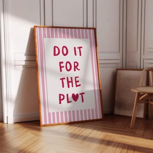 May include: A framed print with the text "DO IT FOR THE PLOT" in red letters. The print has a white background with a pink border and a wooden frame. The print is leaning against a white wall.