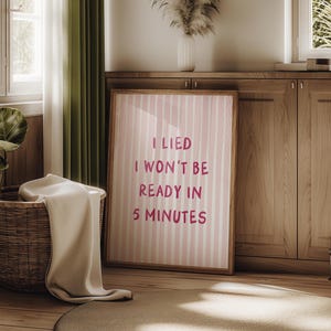 Può includere: Una stampa incorniciata con uno sfondo a righe rosa e bianche e il testo "I LIED I WON'T BE READY IN 5 MINUTES" in grassetto, rosa. La stampa è in una cornice di legno e si appoggia a un mobile di legno.