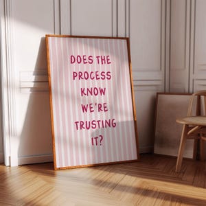 Op de afbeelding: Een ingelijste print met een roze en witte gestreepte achtergrond en de tekst "DOES THE PROCESS KNOW WE'RE TRUSTING IT?" in roze. De lijst heeft een warme bruine kleur en de print leunt tegen een witte muur.