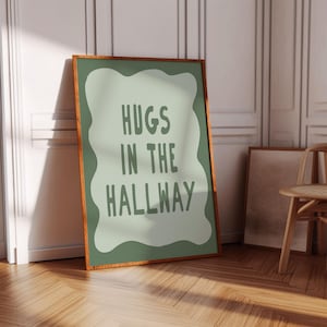 Könnte beinhalten: Ein gerahmter Kunstdruck mit dem Text "HUGS IN THE HALLWAY" in grünen Buchstaben. Das Kunstwerk hat einen hellgrünen Hintergrund mit einer wellenförmigen, unregelmäßigen Form. Der Rahmen ist braun, und der Druck lehnt an einer Wand.