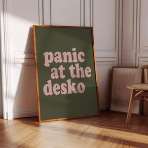 May include: A rectangular wall art print in a wooden frame featuring a dark olive green background with the text "panic at the desko" written in a light pink, rounded, wavy font. The print is positioned upright on a wooden floor surface.
