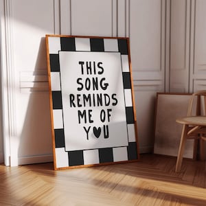 May include: A framed print with the text "THIS SONG REMINDS ME OF YOU" in black on a white background. The frame has a black and white checkered border and a brown wooden frame. The print is leaning against a wall.