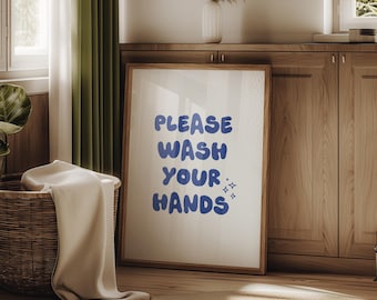 May include: A framed print with the text "PLEASE WASH YOUR HANDS" in blue bubble letters. The artwork is displayed in a room with a woven basket, a beige rug, and wooden cabinets. The print has a wooden frame.
