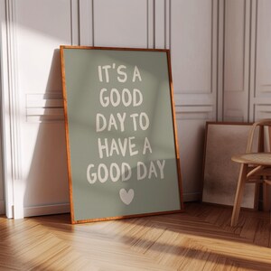 以下が含まれることがあります： セージグリーンの背景に「IT'S A GOOD DAY TO HAVE A GOOD DAY」というテキストが白で書かれたフレーム付きポスター。フレームは温かみのある茶色です。下部には白いハートがあります。ポスターは白い壁に立てかけられています。