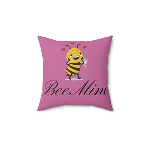 May include: Square throw pillow with a pink background featuring a cartoon bee with a yellow body and black stripes, red hearts, and the words "Bee Mine" in black script.