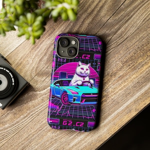Op de afbeelding: Een zwarte telefoonhoes met een witte kat die een blauwe sportwagen bestuurt. De achtergrond is een retro-ontwerp met roze en paarse accenten. De tekst "G2.CZ" is op de hoes gedrukt.