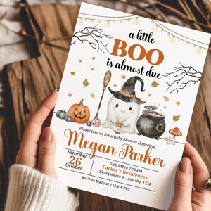 May include: A Halloween-themed baby shower invitation with the text "a little BOO is almost due." The invitation features a ghost wearing a witch hat, a pumpkin, and a cauldron. The invitation is held by a person.