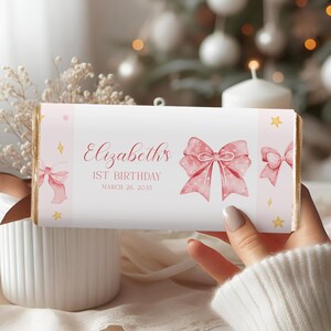 May include: A candy bar wrapper with a pink bow design and the text "Elizabeth's 1st Birthday March 26, 2035". The wrapper has a light pink background with illustrations of bows and stars. The edges of the bar are gold foil.