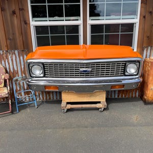 May include: An orange vintage truck front end with a chrome grill and headlights. The front end is mounted on a wooden cart. The background includes rustic metal and wooden elements, and vintage chairs.