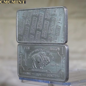 May include: Two silver bullion bars. The top bar has a repeating pattern of "CMCMINT". The bottom bar features a buffalo, the text "UNITED STATES OF AMERICA", and "ONE TROY OUNCE". The bars are marked as .999 fine.