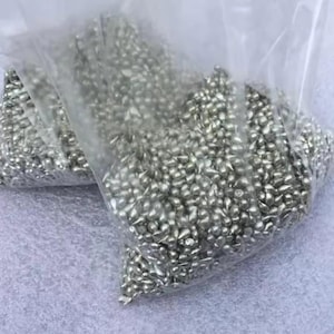 May include: Two clear plastic bags filled with small, silver-colored metal beads. The beads have a teardrop shape and are densely packed within the bags. The bags are slightly transparent, revealing the contents.