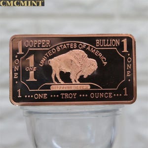 May include: A copper bullion bar with a detailed image of a bison. The bar is rectangular and features the text "1 Copper Bullion One Troy Ounce United States of America".