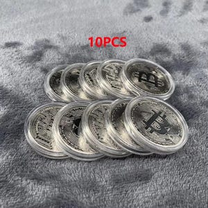 May include: Ten silver-colored Bitcoin coins in clear protective cases are arranged on a gray, textured surface. The coins feature the Bitcoin logo and intricate designs. The text "10PCS" is in red.