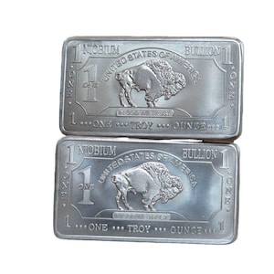 Peut inclure: Deux lingots de niobium argentés. Chaque barre présente une image de bison, le texte "United States of America" et "One Troy Ounce". Les barres sont rectangulaires avec des coins arrondis et le texte "Niobium" en haut.