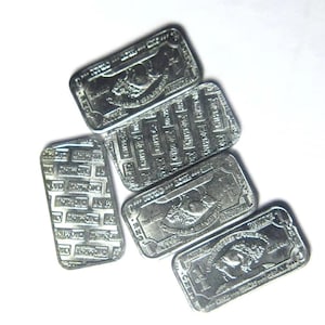 May include: Several rectangular silver bullion bars are displayed. Each bar has detailed engravings, including text and a central image. The bars are arranged on a white surface, highlighting their metallic sheen and intricate designs.