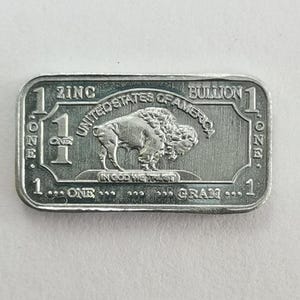 Op de afbeelding: Een rechthoekige zinken bullion munt met een gedetailleerde afbeelding van een bizon. De munt is gemarkeerd met "UNITED STATES OF AMERICA", "ONE GRAM" en "IN GOD WE TRUST". De munt heeft ook de tekst "ZINC" en "BULLION".